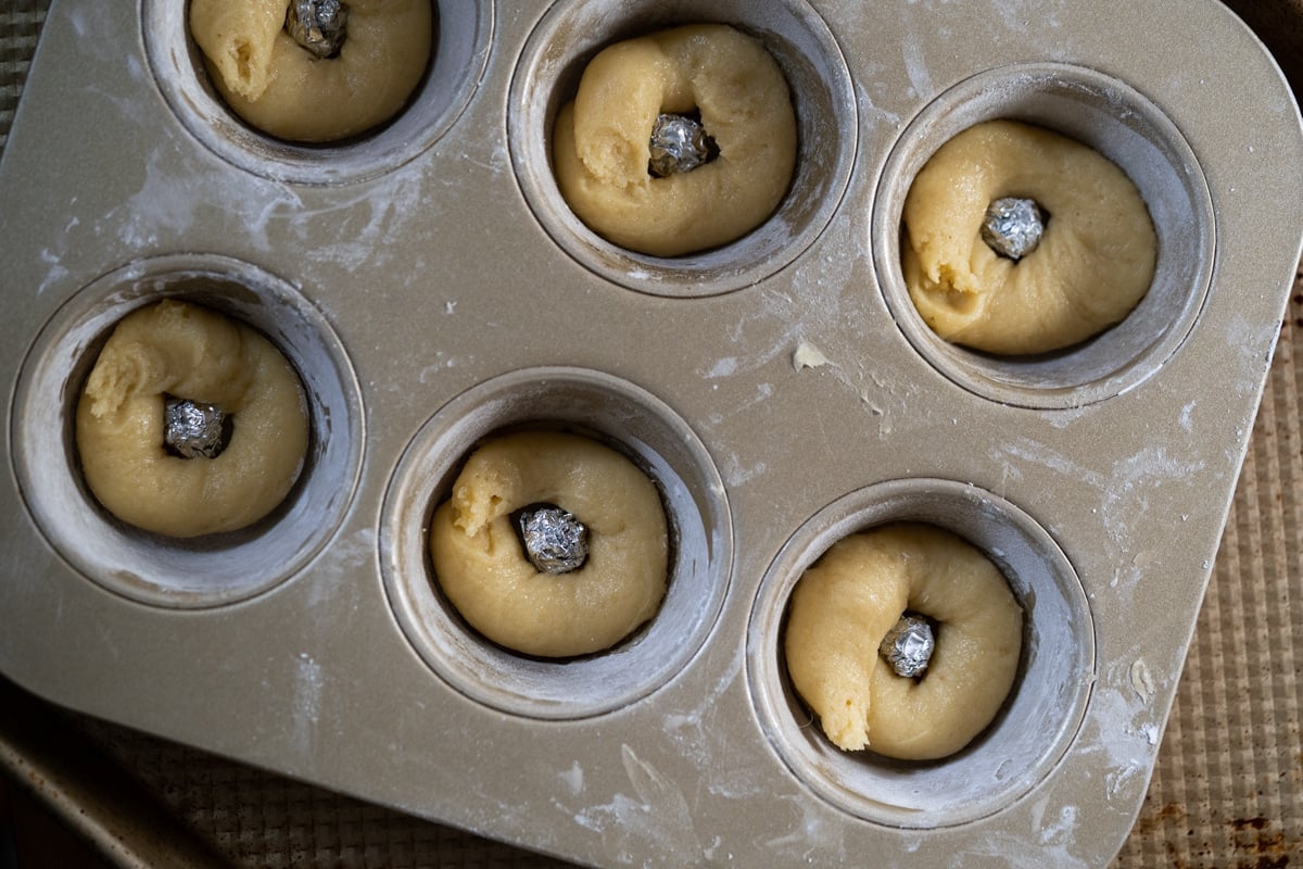 Showing how to make a makeshift donut pan by placing aluminum balls in the middle of muffin pan and piping the batter around it