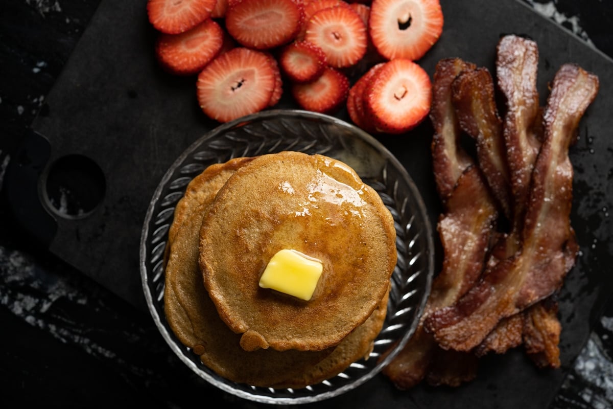 Overhead shot of stacked flourless keto pancakes with sides of strawberries and bacon