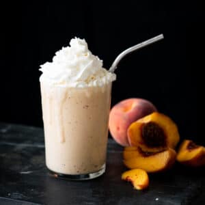 A keto peach milkshake with whipped cream, a metal straw and halved peaches on the side