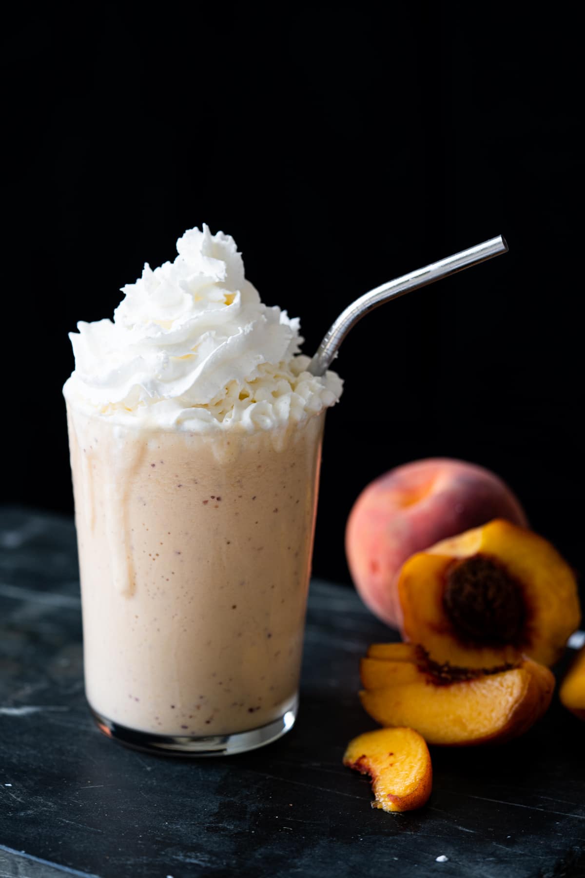 A keto peach milkshake with whipped cream, a metal straw and halved peaches on the side