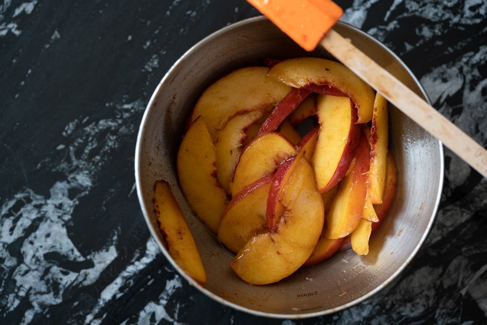 Sliced peaches macerating in a metal bowl over a black marble surface with a spatula on the side